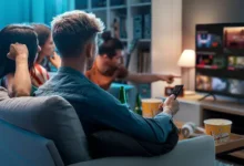 Filmes da Netflix para assistir em família: Grupo de amigos reunidos em uma sala de estar moderna, escolhendo algo para assistir na TV. Um deles segura o controle remoto enquanto outro aponta para a tela, que exibe várias opções de conteúdo. Há pipoca e garrafas de bebida na mesa, criando um clima descontraído e social. O ambiente é iluminado de forma acolhedora, com uma estante ao fundo decorada com livros e objetos. Crédito: Demaerre.