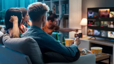 Filmes da Netflix para assistir em família: Grupo de amigos reunidos em uma sala de estar moderna, escolhendo algo para assistir na TV. Um deles segura o controle remoto enquanto outro aponta para a tela, que exibe várias opções de conteúdo. Há pipoca e garrafas de bebida na mesa, criando um clima descontraído e social. O ambiente é iluminado de forma acolhedora, com uma estante ao fundo decorada com livros e objetos. Crédito: Demaerre.