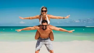 Lugares para passar lua de mel: A imagem mostra um casal sorridente em uma praia paradisíaca, com o homem carregando a mulher sobre os ombros. Ambos estão de braços abertos, simulando asas, em uma pose divertida e descontraída. O cenário ao fundo apresenta mar turquesa, céu azul claro e areia branca, transmitindo uma sensação de alegria, liberdade e clima tropical. Crédito: Pixabay.