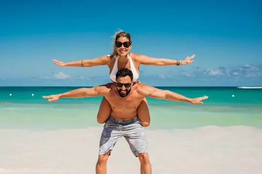 Lugares para passar lua de mel: A imagem mostra um casal sorridente em uma praia paradisíaca, com o homem carregando a mulher sobre os ombros. Ambos estão de braços abertos, simulando asas, em uma pose divertida e descontraída. O cenário ao fundo apresenta mar turquesa, céu azul claro e areia branca, transmitindo uma sensação de alegria, liberdade e clima tropical. Crédito: Pixabay.