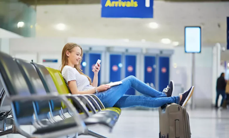 Km de Vantagens: Jovem mulher relaxa em aeroporto enquanto troca pontos do Km de Vantagens por milhas no celular, planejando sua próxima viagem com bônus de até 60% no LATAM Pass.