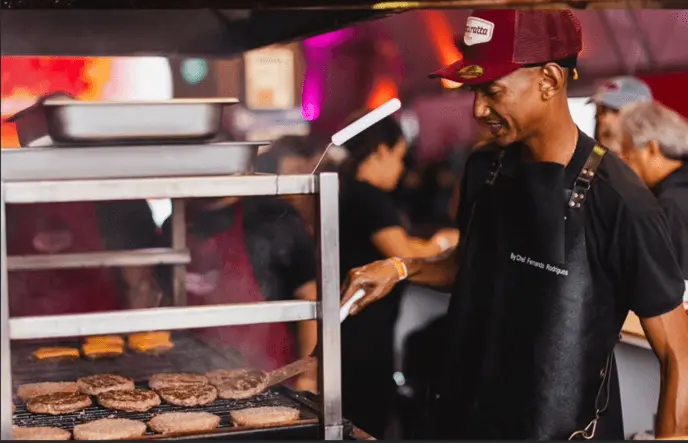 sp-gastronomia-sabor-em-2025 SP GASTRONOMIA 2025: A imagem mostra um chef sorridente — vestindo avental preto e boné vermelho da marca Maturatta — manuseia os hambúrgueres em um ambiente animado e iluminado, típico de eventos gastronômicos. O cenário sugere uma atmosfera descontraída e calorosa, com o público ao fundo, reforçando o clima de festival. Crédito: Divulgação.