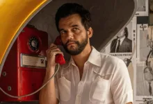 Wagner Moura em cena do filme O Agente Secreto, usando telefone público em ambiente urbano.