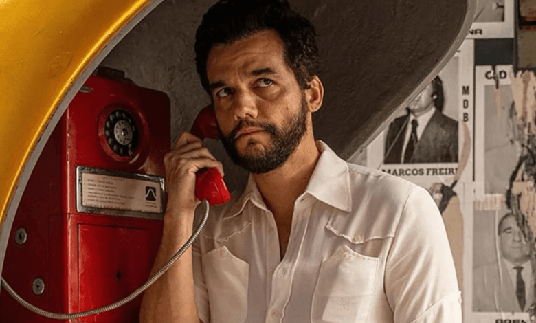 Wagner Moura em cena do filme O Agente Secreto, usando telefone público em ambiente urbano.