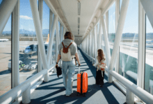 Pessoa adulta e criança caminham por uma ponte de embarque em aeroporto, puxando uma mala de viagem.