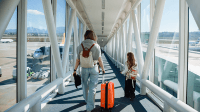 Pessoa adulta e criança caminham por uma ponte de embarque em aeroporto, puxando uma mala de viagem.