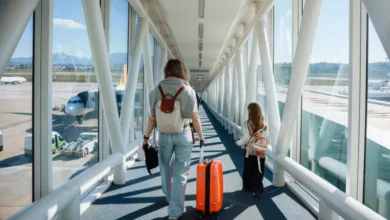 Pessoa adulta e criança caminham por uma ponte de embarque em aeroporto, puxando uma mala de viagem.