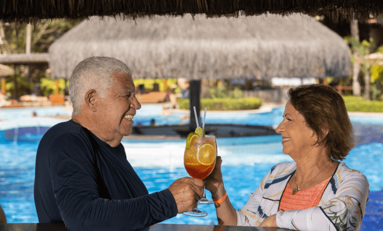 Casal de idosos brinda com drinques em bar dentro da piscina de um resort.