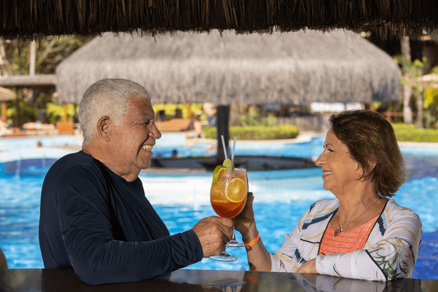 Casal de idosos brinda com drinques em bar dentro da piscina de um resort.