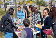 Pessoas reunidas ao redor de uma mesa escolhem livros durante feira de doação em área externa.