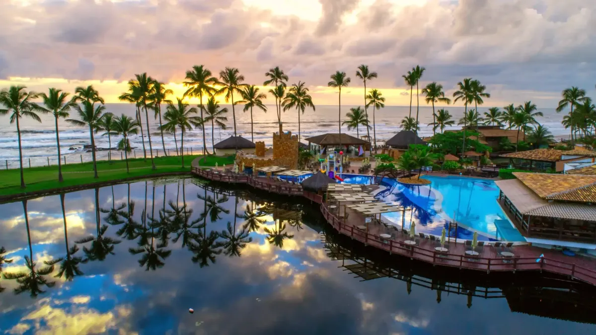 Vista aérea de resort à beira-mar com piscinas, coqueiros e passarela de madeira ao pôr do sol.