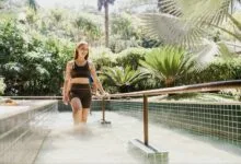 Mulher caminha em piscina de hidroterapia ao ar livre, cercada por vegetação, em ambiente de spa.