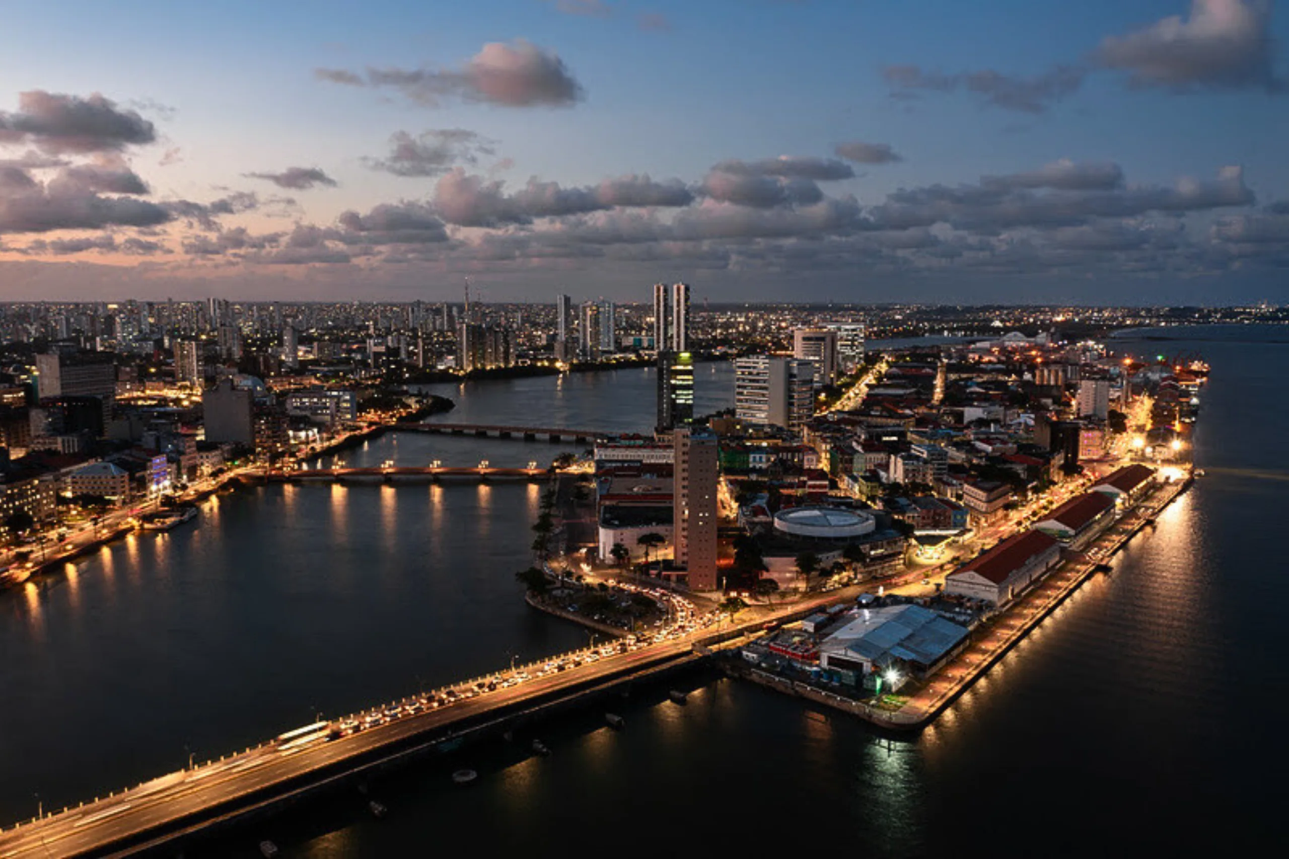 Vista aérea do Recife ao entardecer, com rios, pontes e prédios iluminados.