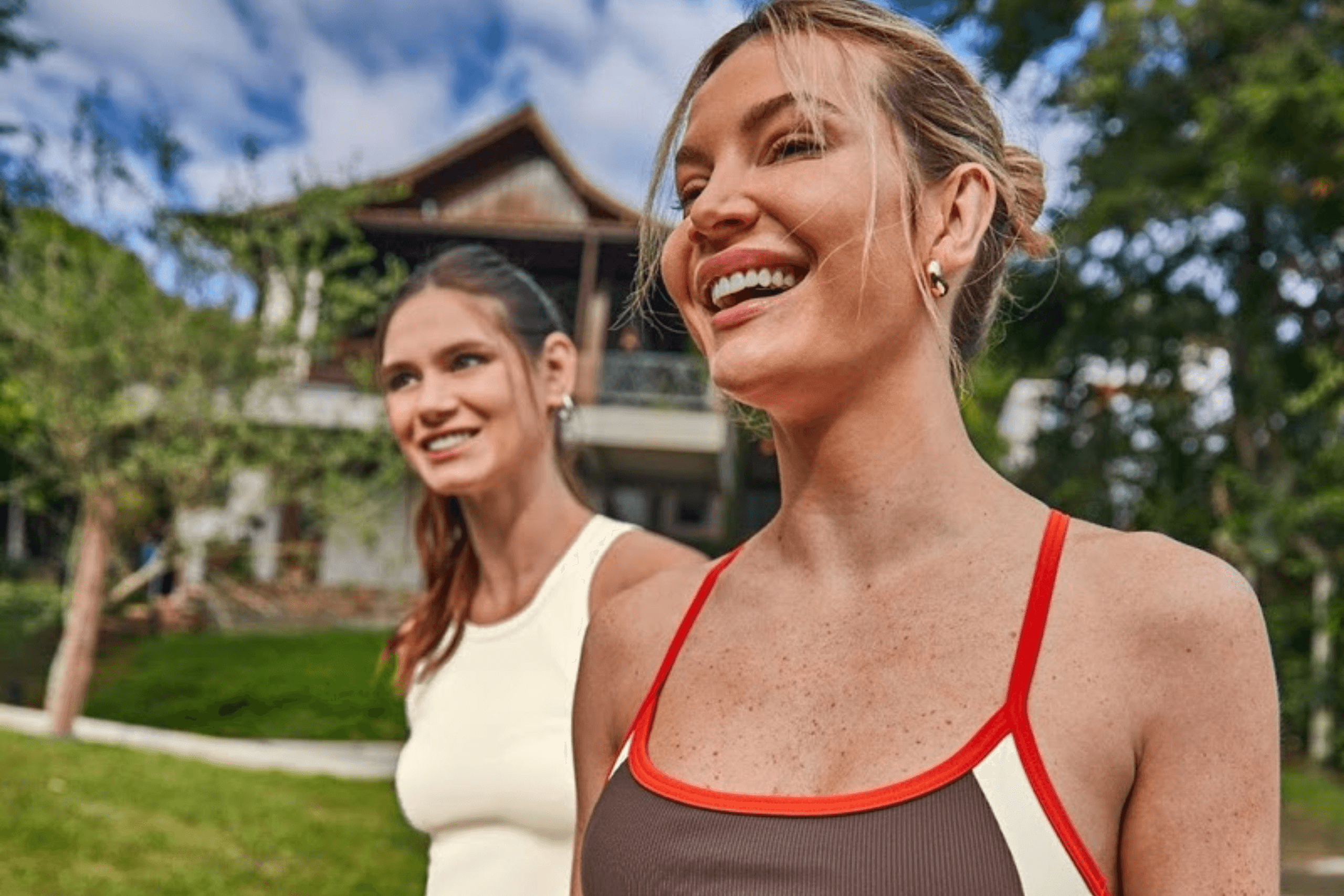 Duas mulheres sorridentes ao ar livre vestindo roupas da Linha Active da Malwee. Uma usa um top marrom com detalhes em laranja e a outra uma regata bege, ideal para passeios de verão.