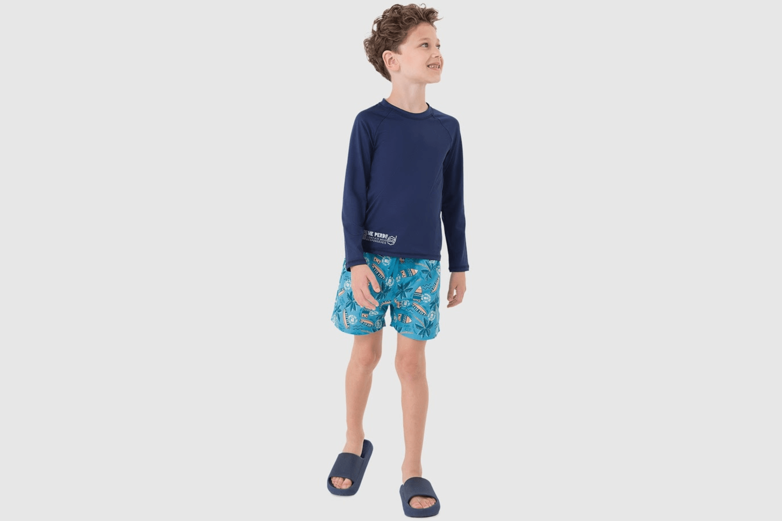 Menino sorridente vestindo conjunto de praia da Malwee Kids: camisa de manga longa azul escura com proteção UV e bermuda azul clara estampada com elementos de surfe.