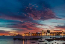 Vista panorâmica da linha do horizonte de Montevidéu ao crepúsculo. O céu exibe nuvens em tons de roxo, rosa e laranja sobre as luzes acesas da cidade e o Rio da Prata.