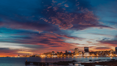 Vista panorâmica da linha do horizonte de Montevidéu ao crepúsculo. O céu exibe nuvens em tons de roxo, rosa e laranja sobre as luzes acesas da cidade e o Rio da Prata.