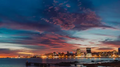 Vista panorâmica da linha do horizonte de Montevidéu ao crepúsculo. O céu exibe nuvens em tons de roxo, rosa e laranja sobre as luzes acesas da cidade e o Rio da Prata.