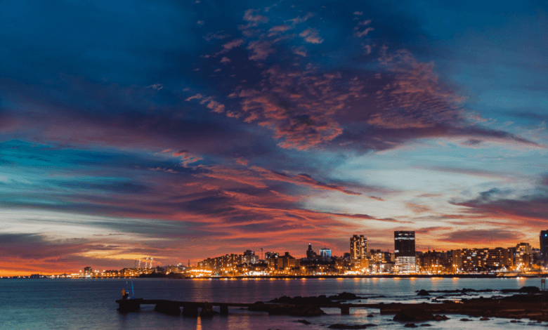 Vista panorâmica da linha do horizonte de Montevidéu ao crepúsculo. O céu exibe nuvens em tons de roxo, rosa e laranja sobre as luzes acesas da cidade e o Rio da Prata.