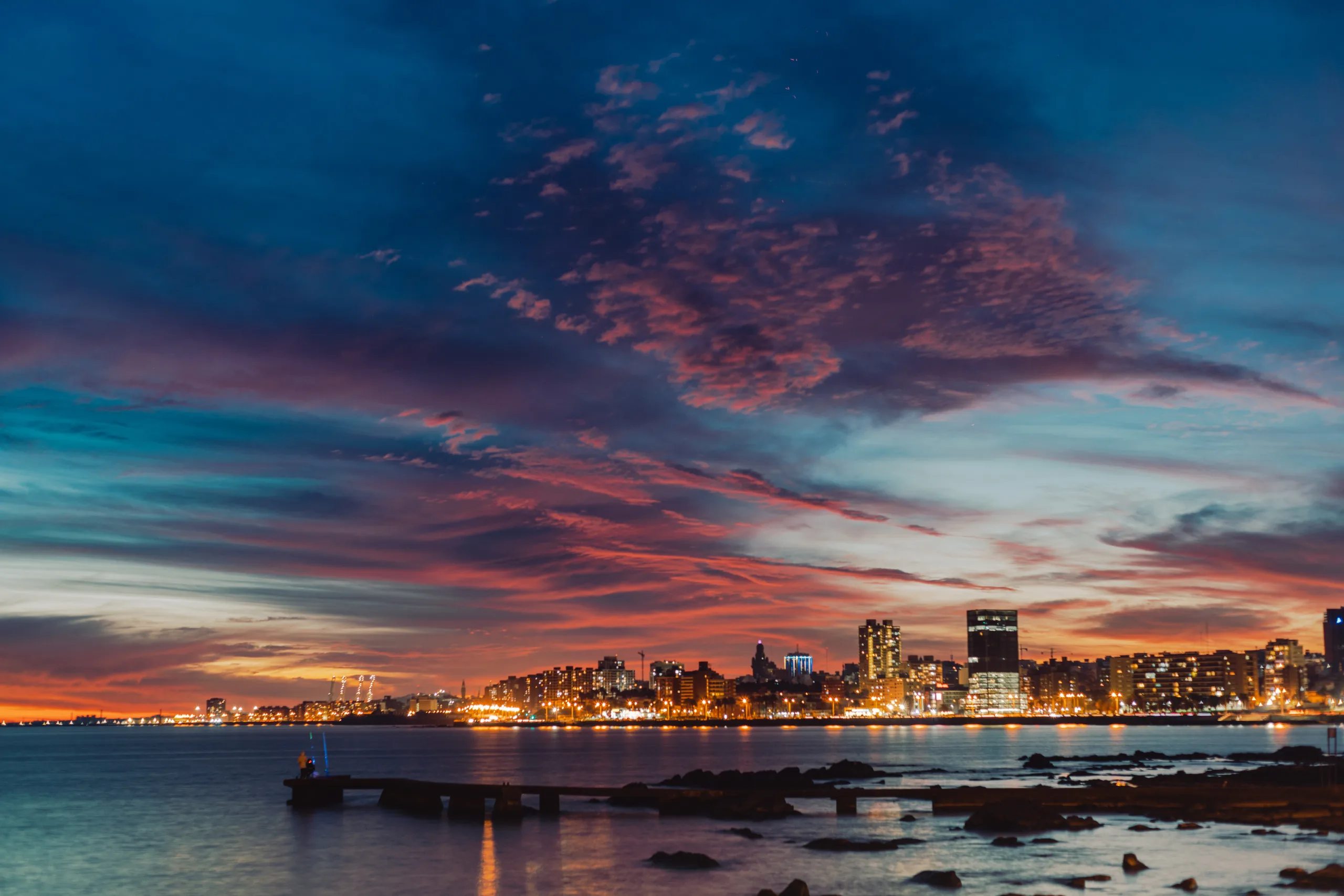 Vista panorâmica da linha do horizonte de Montevidéu ao crepúsculo. O céu exibe nuvens em tons de roxo, rosa e laranja sobre as luzes acesas da cidade e o Rio da Prata.