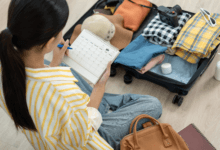 Mulher sentada no chão organizando uma mala de viagem aberta. Ela segura um calendário de planejamento enquanto seleciona roupas dobradas, chapéu e câmera fotográfica.