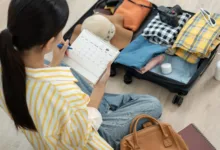 Mulher sentada no chão organizando uma mala de viagem aberta. Ela segura um calendário de planejamento enquanto seleciona roupas dobradas, chapéu e câmera fotográfica.