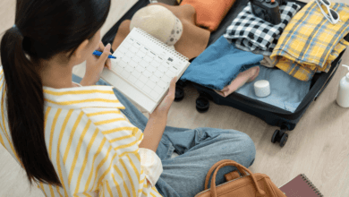 Mulher sentada no chão organizando uma mala de viagem aberta. Ela segura um calendário de planejamento enquanto seleciona roupas dobradas, chapéu e câmera fotográfica.