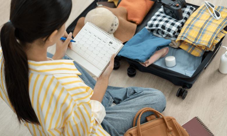 Mulher sentada no chão organizando uma mala de viagem aberta. Ela segura um calendário de planejamento enquanto seleciona roupas dobradas, chapéu e câmera fotográfica.