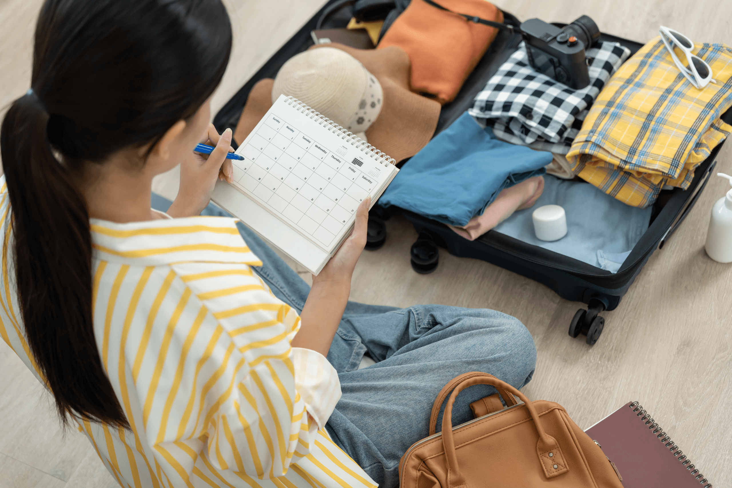 Mulher sentada no chão organizando uma mala de viagem aberta. Ela segura um calendário de planejamento enquanto seleciona roupas dobradas, chapéu e câmera fotográfica.