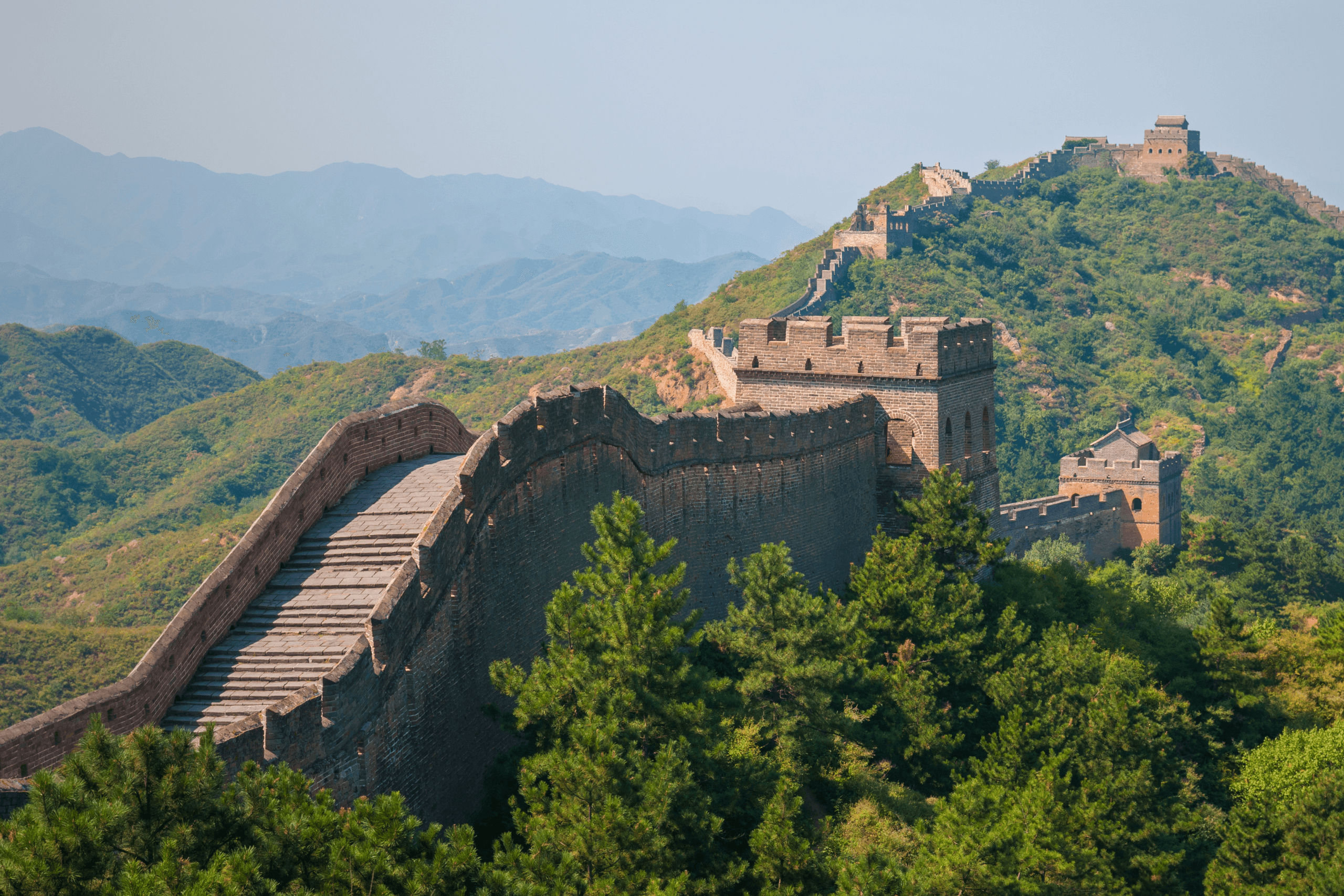 Vista panorâmica da Grande Muralha da China em um dia ensolarado, cercada por montanhas arborizadas sob um céu azul claro.