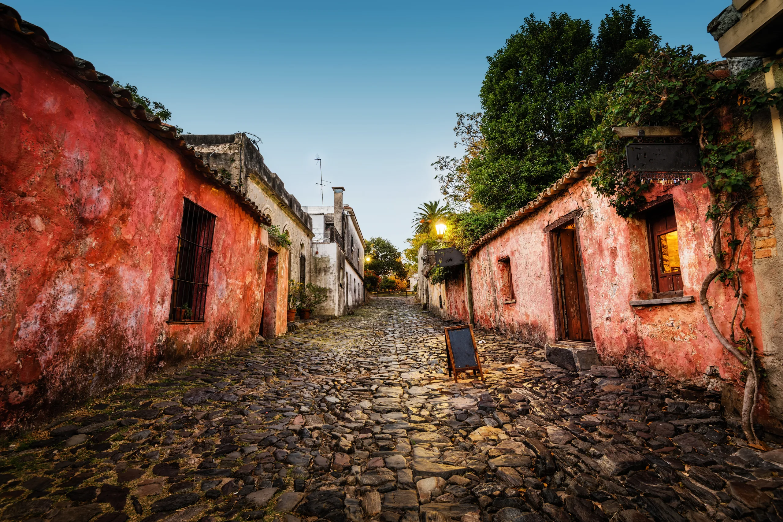 Fotografia de uma rua estreita de paralelepípedos em Colonia del Sacramento, ladeada por casas coloniais de paredes rústicas em tons de rosa e laranja sob um céu de fim de tarde.