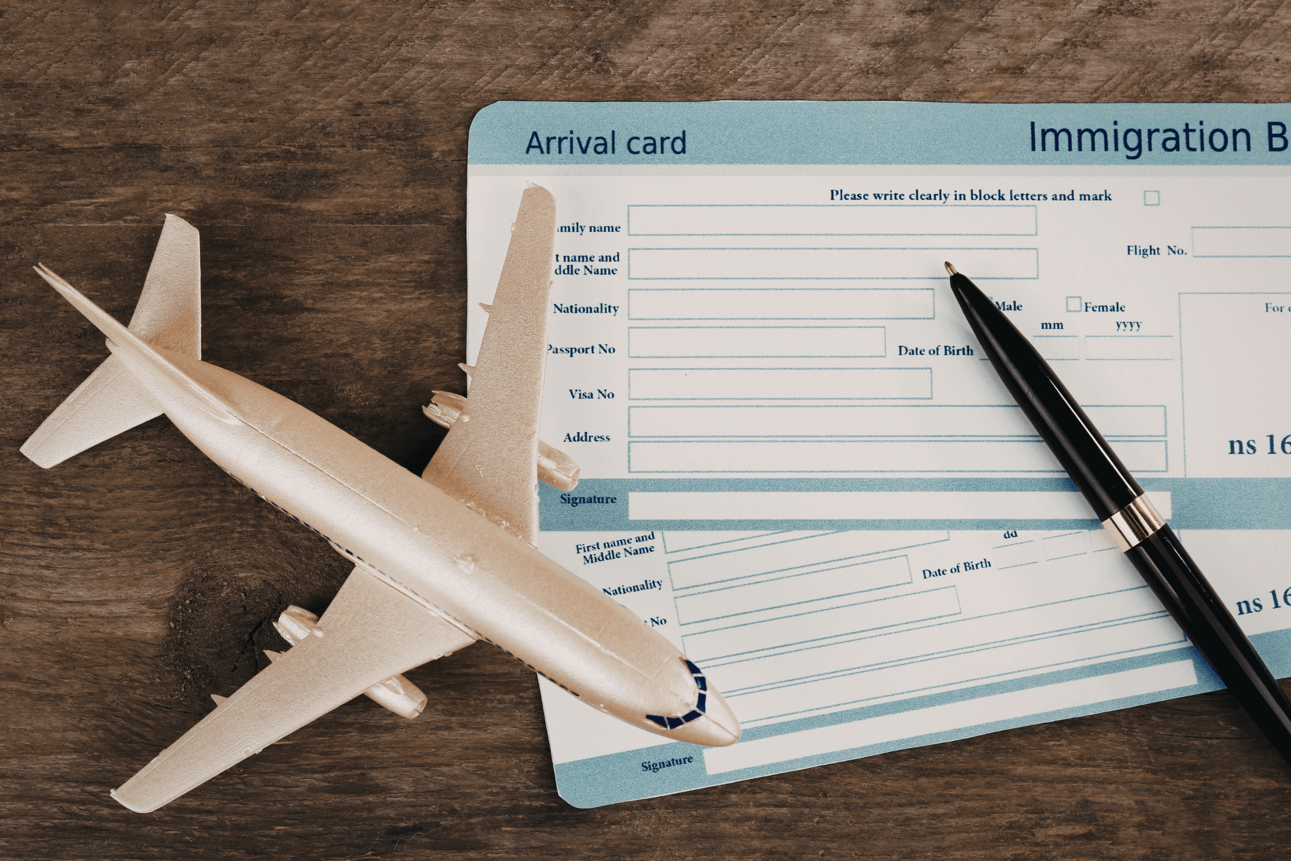 Formulário de imigração azul e branco sobre mesa de madeira, acompanhado de uma caneta e uma miniatura de avião.