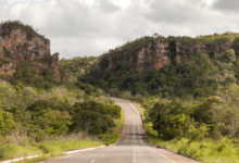 Uma estrada de asfalto cinza com faixas amarelas centrais atravessando um vale verde cercado por grandes paredões de rocha e morros cobertos de árvores.
