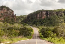 Uma estrada de asfalto cinza com faixas amarelas centrais atravessando um vale verde cercado por grandes paredões de rocha e morros cobertos de árvores.
