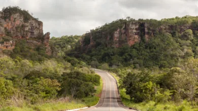 Uma estrada de asfalto cinza com faixas amarelas centrais atravessando um vale verde cercado por grandes paredões de rocha e morros cobertos de árvores.