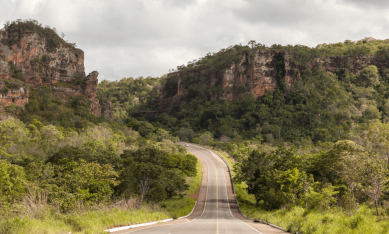 Uma estrada de asfalto cinza com faixas amarelas centrais atravessando um vale verde cercado por grandes paredões de rocha e morros cobertos de árvores.