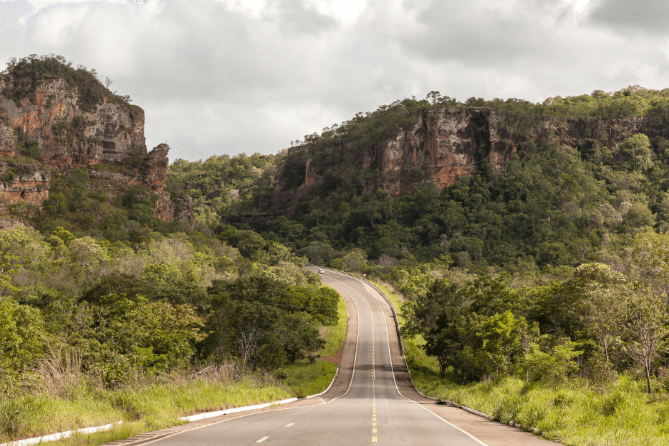Uma estrada de asfalto cinza com faixas amarelas centrais atravessando um vale verde cercado por grandes paredões de rocha e morros cobertos de árvores.
