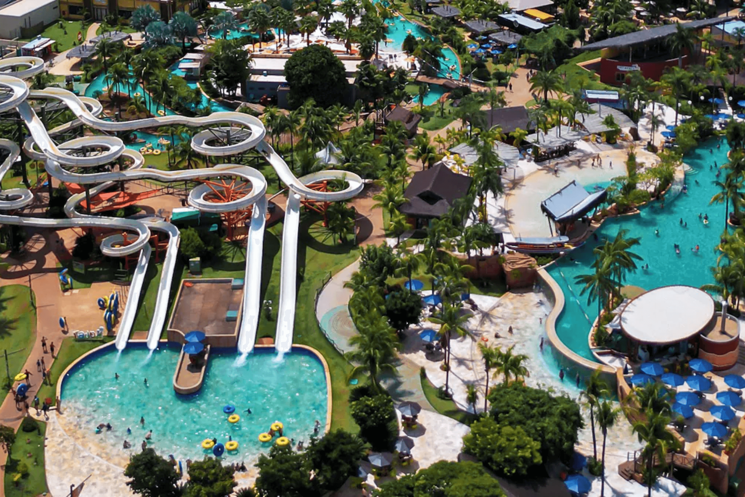 Vista aérea de um grande parque aquático com diversos toboáguas brancos sinuosos, piscinas de águas azuis claras e áreas arborizadas com palmeiras.