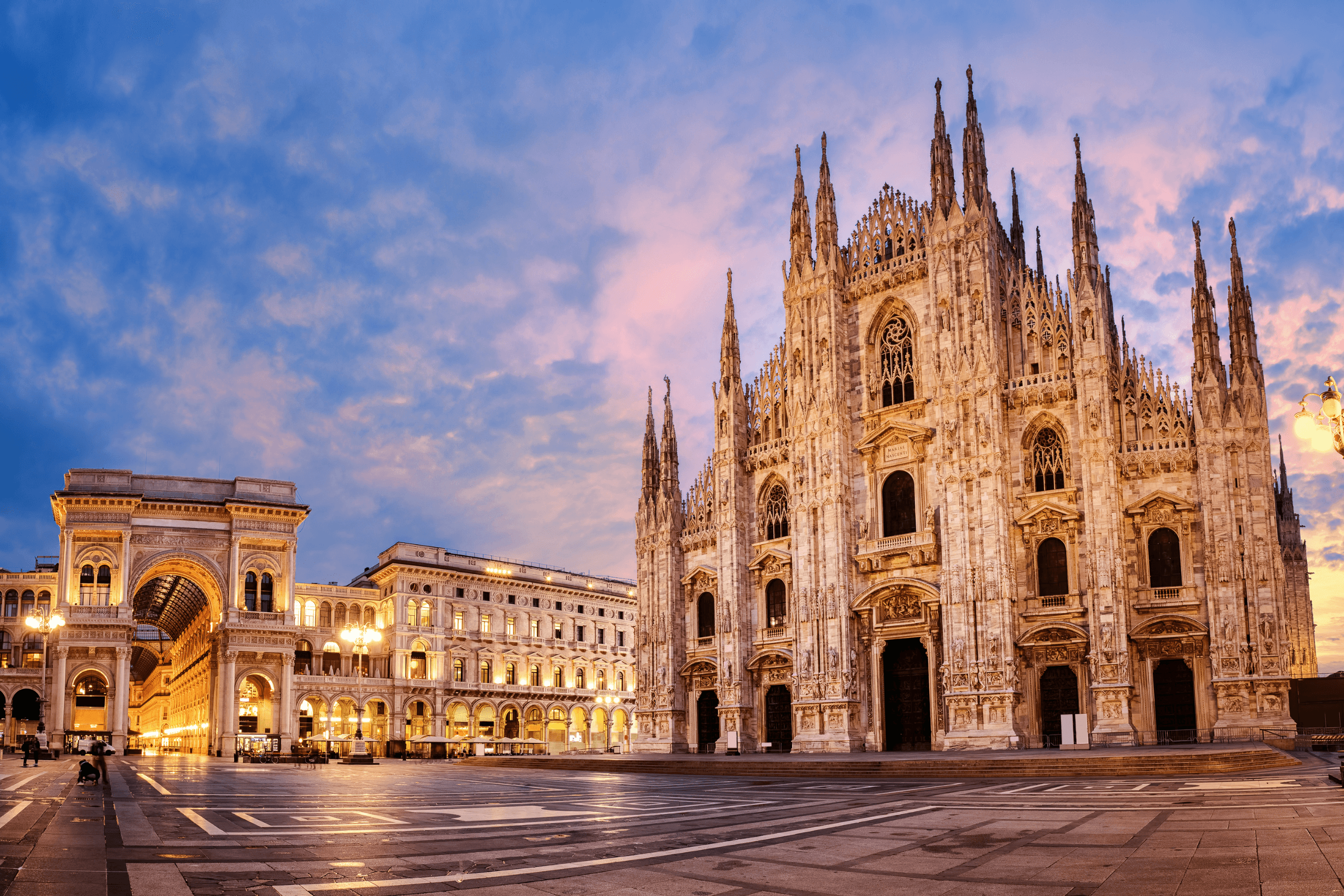 Fachada detalhada da Catedral de Milão (Duomo) e a entrada da Galeria Vittorio Emanuele II sob um céu de entardecer em tons de rosa e azul.