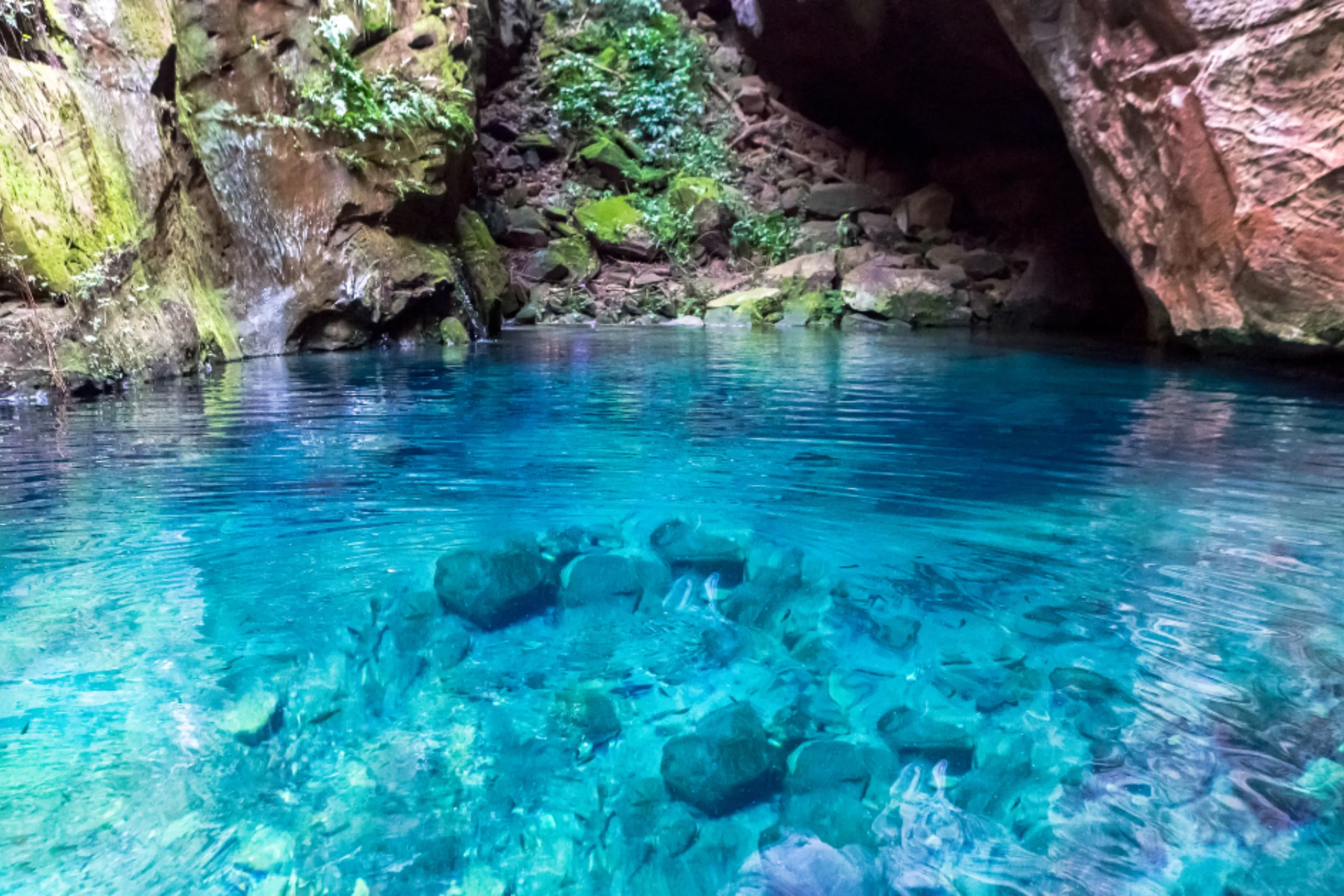 Uma piscina natural de águas azuis extremamente transparentes dentro de uma gruta rochosa, permitindo ver nitidamente as pedras no fundo.