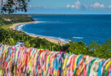 Mirante em Porto Seguro com vista para o mar. Em primeiro plano, uma grade repleta de fitinhas coloridas do Senhor do Bonfim. Ao fundo, a orla marítima com areia clara, coqueirais e mar azul degradê.