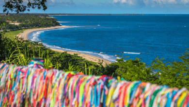 Mirante em Porto Seguro com vista para o mar. Em primeiro plano, uma grade repleta de fitinhas coloridas do Senhor do Bonfim. Ao fundo, a orla marítima com areia clara, coqueirais e mar azul degradê.