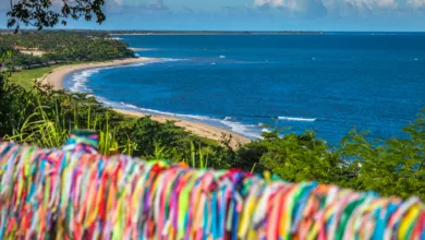 Mirante em Porto Seguro com vista para o mar. Em primeiro plano, uma grade repleta de fitinhas coloridas do Senhor do Bonfim. Ao fundo, a orla marítima com areia clara, coqueirais e mar azul degradê.