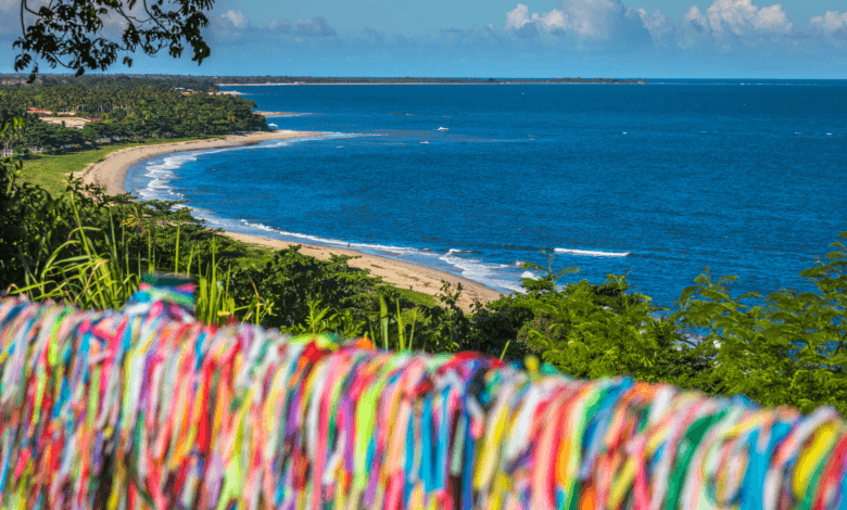 Mirante em Porto Seguro com vista para o mar. Em primeiro plano, uma grade repleta de fitinhas coloridas do Senhor do Bonfim. Ao fundo, a orla marítima com areia clara, coqueirais e mar azul degradê.