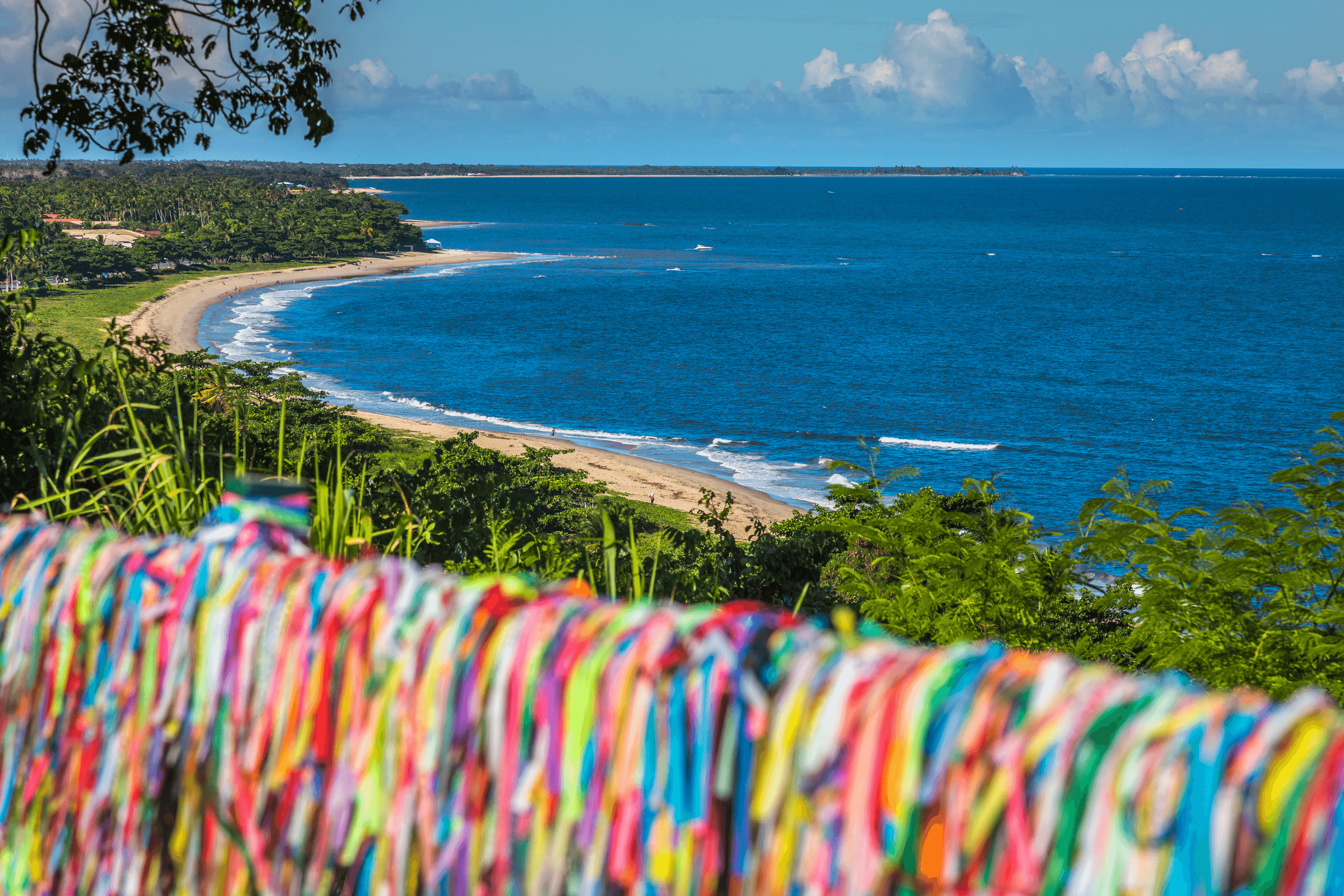 Mirante em Porto Seguro com vista para o mar. Em primeiro plano, uma grade repleta de fitinhas coloridas do Senhor do Bonfim. Ao fundo, a orla marítima com areia clara, coqueirais e mar azul degradê.