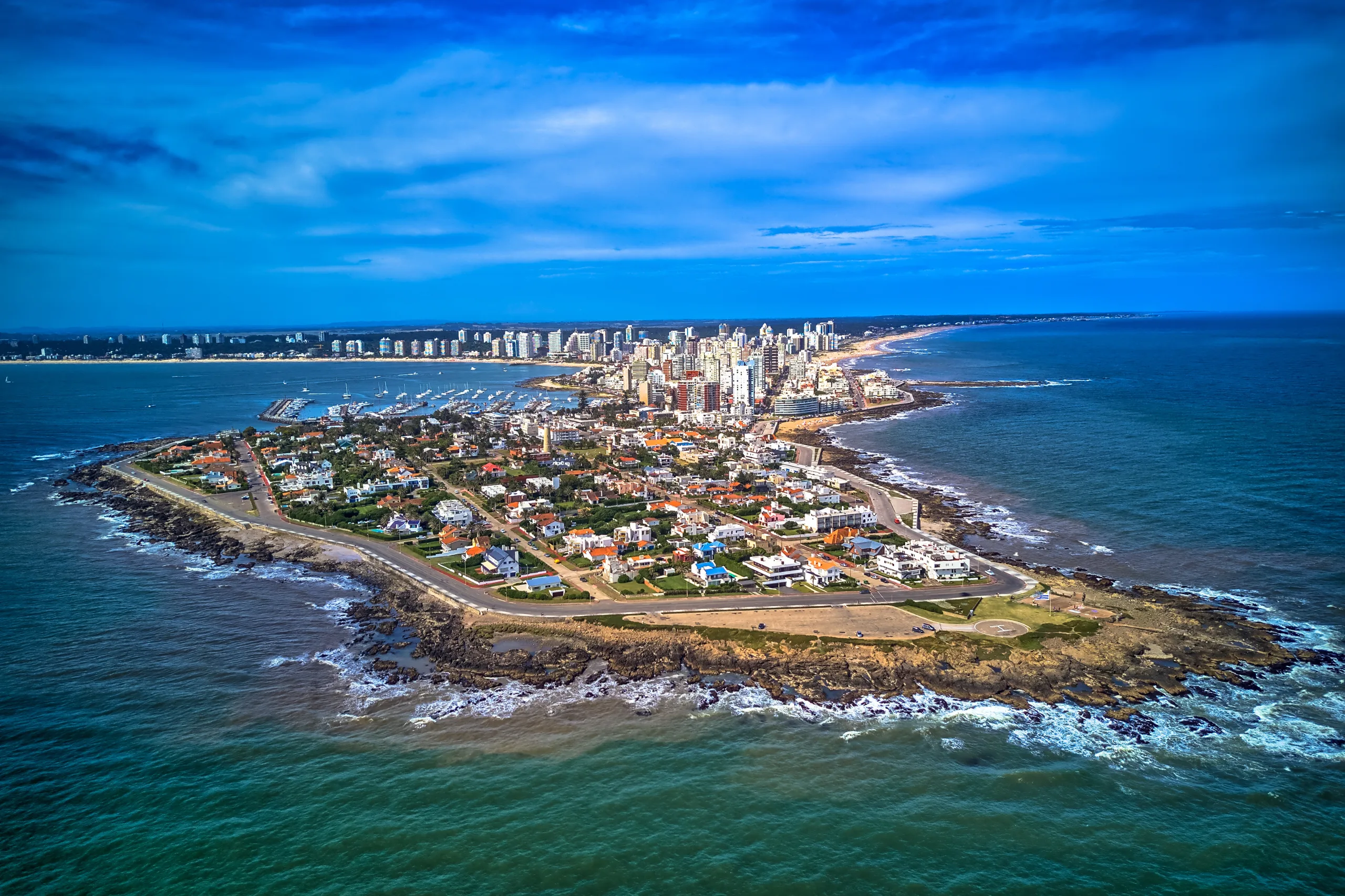 Fotografia aérea da península de Punta del Este, mostrando a densa concentração de edifícios, praias circundantes e a marina repleta de barcos.