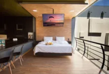 Quarto de motel moderno com cama de casal branca, painéis de madeira no teto e na parede, e uma grande janela de vidro à direita.