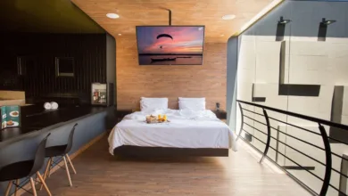 Quarto de motel moderno com cama de casal branca, painéis de madeira no teto e na parede, e uma grande janela de vidro à direita.