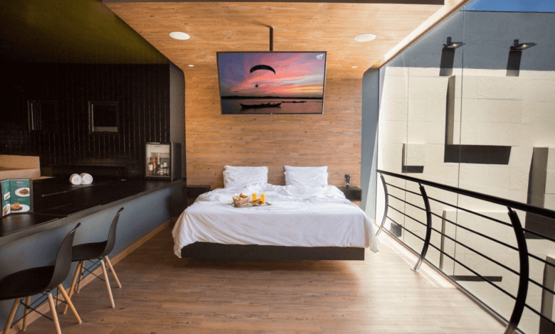 Quarto de motel moderno com cama de casal branca, painéis de madeira no teto e na parede, e uma grande janela de vidro à direita.