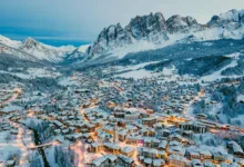 Vista aérea da vila alpina de Cortina d'Ampezzo coberta de neve ao anoitecer, com as montanhas Dolomitas ao fundo e luzes urbanas acesas.
